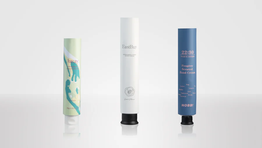 Slim aluminum hand cream tubes in pale green, white, and blue printed finishes, shown standing on white and black flip-top dispensing caps.