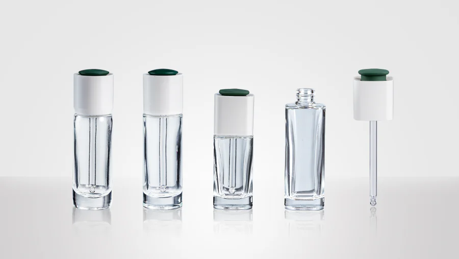 Clear rotary dropper bottles in tall cylindrical and compact faceted shapes with white closures and dark green push tops, shown alongside an open bottle and separate glass pipette assembly.